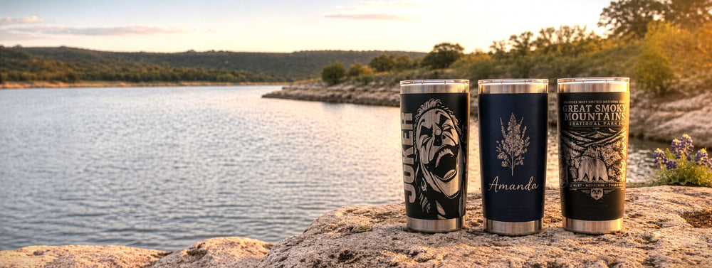 Three tumblers with custom artwork and name engraving beside a lake at sunset.